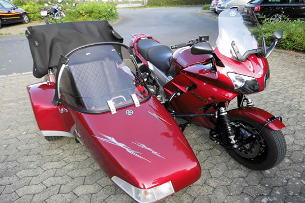 Ein rotes Motorrad mit einem angeschlossenen Beiwagen. Das Motorrad verfügt über eine Windschutzscheibe und ist auf einer gepflasterten Oberfläche geparkt, möglicherweise auf einem Parkplatz. Der Beiwagen hat ebenfalls eine eigene Windschutzscheibe und scheint mit einem schwarzen Verdeck bedeckt zu sein. Sowohl das Motorrad als auch der Beiwagen haben weiße Streifen oder Aufkleber entlang ihrer Karosserien. Dieses Bild könnte für Motorrad- oder Beiwagen-Enthusiasten oder für diejenigen interessant sein, die einzigartige Fahrzeugmodifikationen schätzen.